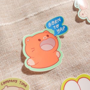 May include: A collection of colorful stickers on a tan fabric surface. One sticker features an orange cartoon cat with the text "Born to Yap" in a blue speech bubble. Other stickers with cartoon animals are also visible.