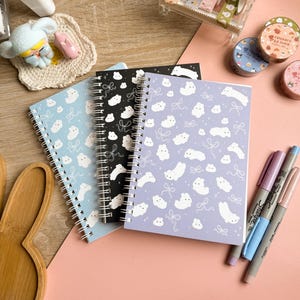Könnte beinhalten: Drei spiralgebundene Notizbücher in Hellblau, Schwarz und Lila mit einem sich wiederholenden Muster aus weißen Cartoon-Tieren und Schleifen. Die Notizbücher liegen auf einer rosa Oberfläche, zusammen mit bunten Markern und dekorativem Klebeband.