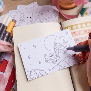 May include: A person colouring a black and white illustration of a bunny in a sleigh with reindeer under a full moon. The illustration is on a white card, with a black notebook and markers nearby. Pink ribbon and decorative tape are also visible.