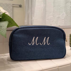 May include: A denim blue cosmetic bag with a gold zipper and the embroidered initials "MM" in white. The bag is sitting on a white tiled surface, with a white vase and a green candle holder in the background.