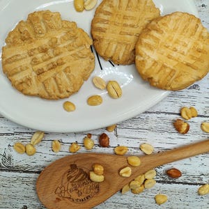 Puede incluir: Tres galletas de mantequilla de cacahuete en un plato blanco con cacahuetes esparcidos alrededor del plato. El plato está sobre una superficie de madera. Una cuchara de madera con el texto "Heavens to Betsy Cakery" está en la esquina inferior derecha de la imagen.