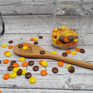 May include: A wooden spoon with a brown and yellow design on a white wooden surface. There are brown, yellow, and orange candies scattered around the spoon. A glass jar filled with the same candies is in the background.