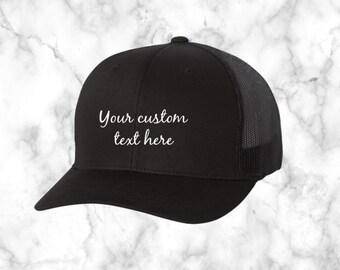 May include: Black trucker hat with a black mesh back. The hat has white embroidery that says "Your custom text here".