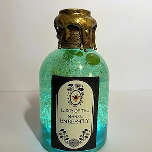 Op de afbeelding: Een decoratieve glazen fles gevuld met een turquoise vloeistof en glitter. De fles heeft een goudkleurige dop met een druipwas-effect. Een zwart etiket met witte tekst luidt "Elixir of the Marsh Ember-Fly".