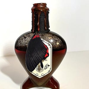 Op de afbeelding: Een hartvormige, donkerbruine fles met een zwarte en rode vlindervleugelbedel. De fles heeft een getextureerde, donkerbruine bovenkant met een zwart touwdetail. Een wit label met tekst en een vlindervleugelontwerp hangt aan de fles.