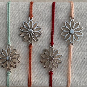 May include: Four silver daisy charm bracelets with different colored cords. The cords are mint green, orange, red, and peach.