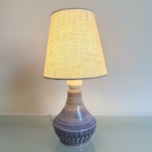 Op de afbeelding: Een keramische tafellamp met een beige stoffen lampenkap. De lampvoet is blauwgrijs van kleur met een textuur, geribbeld ontwerp. De lamp is ingeschakeld en verlicht de kap.