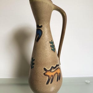 Op de afbeelding: Een bruin keramieken kan met een gebogen handvat en een uitlopende tuit. De kan is versierd met blauwe, groene en oranje abstracte ontwerpen. De kan is ongeveer 25 cm hoog.