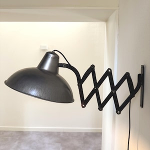 Könnte beinhalten: Eine schwarze Wandleuchte im Industriestil mit einem verstellbaren Scherenarm. Die Lampe hat einen metallgrauen Schirm und ein schwarzes Kabel. Die Lampe ist an einer weißen Wand montiert.