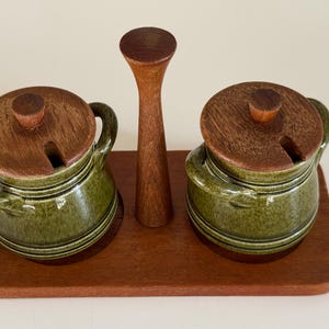 Puede incluir: Un juego de condimentos de madera con dos botes de cerámica verde con tapas y asas de madera, un utensilio central de madera y una bandeja de madera. El juego está diseñado para servir condimentos.