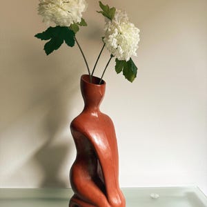 Op de afbeelding: Een terracotta-kleurige vaas in de vorm van een knielende figuur, met twee witte hortensia bloemen met groene bladeren. De vaas is gemaakt van een glad, gepolijst materiaal en staat op een glazen oppervlak.