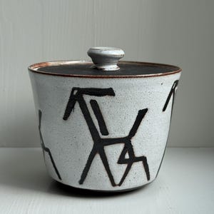 Op de afbeelding: Een keramische pot met deksel, een gespikkelde witte buitenkant en een donkerbruine rand en deksel. De pot heeft abstracte zwarte geometrische ontwerpen. Het deksel heeft een kleine, afgeronde knop. De pot wordt waarschijnlijk gebruikt voor opslag.