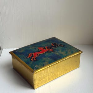 Op de afbeelding: Rechthoekige vintage doos met een gouden geometrisch patroon op de zijkanten. Het deksel is blauwgroen en blauw met een rood en zwart paardenontwerp. De doos heeft gouden randen en is waarschijnlijk van metaal.