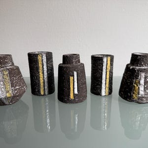 Puede incluir: Cinco jarrones de cerámica marrón oscuro con rayas verticales blancas y doradas. Los jarrones tienen formas variadas, incluyendo diseños cilíndricos y geométricos. Los jarrones están dispuestos sobre una superficie reflectante.