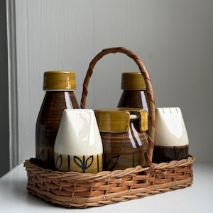 Op de afbeelding: Een vintage keramische kruidenset in een geweven mand. De set bevat twee flessen, een suikerpot en twee zoutvaatjes. De flessen en de suikerpot zijn bruin met gele deksels. De zoutvaatjes zijn wit met zwarte bladmotieven.