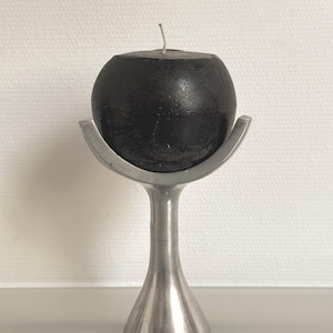May include: A spherical black candle rests inside a silver-colored metal candle holder. The holder features a flared, pedestal-style base that tapers upward into a curved, bowl-shaped top designed to cradle the round candle securely in place.