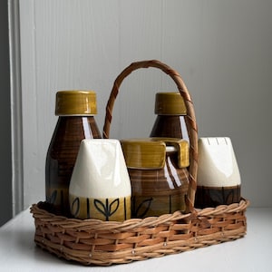Op de afbeelding: Een vintage bruin en geel keramisch condimentenset in een geweven mand. De set bevat twee flessen, een suikerpot en twee strooiers. De keramische stukken hebben een bruin en geel gestreept ontwerp met zwarte bladaccenten.