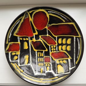 May include: A decorative ceramic plate with a black background and a village scene. The plate features red, yellow, and white accents, depicting buildings and a sun-like shape. The plate has a diameter of approximately 10 inches.