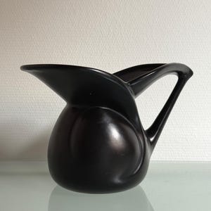 May include: A black ceramic jug with a distinctive, curved design. The jug has a rounded body and a handle that curves upwards. The spout is elongated and pointed. The jug is on a reflective surface.
