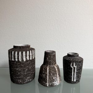 Puede incluir: Tres jarrones de cerámica texturizada en tonos marrones y blancos. Los jarrones tienen forma cilíndrica, cónica y redondeada. Los jarrones tienen líneas blancas verticales y horizontales. Los jarrones están sobre una superficie reflectante.