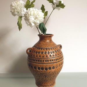Op de afbeelding: Een bruine keramische vaas met een textuuroppervlak en decoratieve zwarte geometrische patronen. De vaas heeft twee handvatten en is gevuld met witte bloemen met groene bladeren. De vaas is ongeveer 30 cm hoog.
