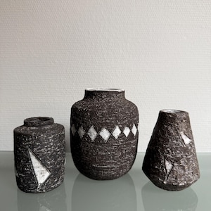 Puede incluir: Tres jarrones de cerámica texturizada en tonos marrón y negro. Los jarrones tienen diseños geométricos blancos. El jarrón de la izquierda es cilíndrico con un diseño triangular. El jarrón central es redondo con un patrón de diamantes. El jarrón de la derecha tiene forma de cono.
