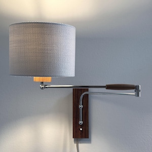 May include: A wall-mounted lamp with a gray cylindrical lampshade. The lamp features a wooden handle and a dark wood base. The metal arm extends from the base, supporting the shade. The lamp is plugged into a wall outlet.
