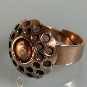 Op de afbeelding: Een metalen ring met een groot, cirkelvormig, koepelvormig middelpunt met een glad, gepolijst koperkleurig centrum, omringd door een getextureerde, geschulpte rand met meerdere donkere, verzonken cirkelvormige inkepingen.