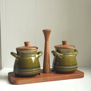 Op de afbeelding: Een houten condimentenset met twee groene keramische potten met deksels en handvatten, geplaatst op een houten dienblad. Een hoog houten stuk staat tussen de potten. De potten hebben horizontale lijnen.