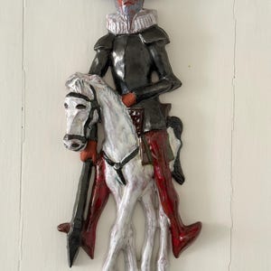 Op de afbeelding: Een keramische wandplaat met een figuur in een zwarte hoed, donker harnas en een witte kraag op een wit paard. De figuur draagt een rode broek en houdt een lange zwarte speer vast. Het object heeft een glanzende afwerking met gedetailleerde texturen.