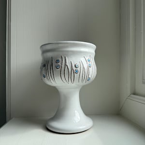 Op de afbeelding: Een witte keramische voetplanter met een brede bovenkant en een uitlopende basis. De planter is versierd met verticale bruine lijnen en kleine blauwe stippen. De planter staat op een wit oppervlak.