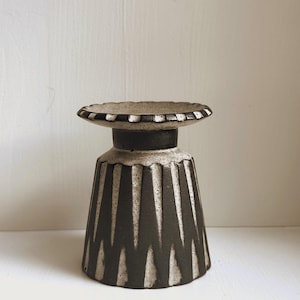 May include: A handcrafted, ceramic pedestal bowl with a dark gray base and a light gray rim. The bowl features a geometric pattern of vertical white lines against the dark gray. The rim has a decorative, dotted design. This unique piece is ideal for displaying small items.