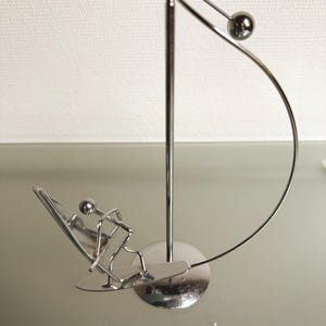 May include: A silver-colored metal sculpture depicting a surfer on a surfboard. The sculpture has a minimalist design with a curved arc and a small ball. The surfer is in a riding position, adding a dynamic element to the piece.