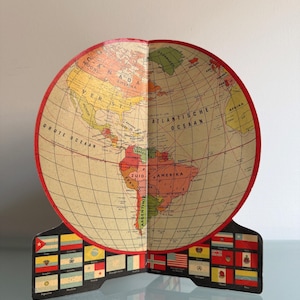 Op de afbeelding: Een vintage papieren wereldbol met een rode rand, met een wereldkaart met continenten en landen. De wereldbol is open, waardoor de binnenkant zichtbaar is, en staat op een voetstuk versierd met diverse vlaggen. Het is een decoratief item.