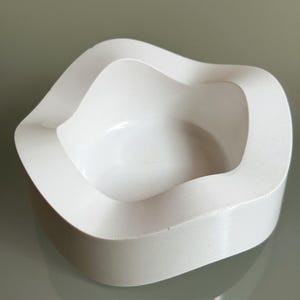 Op de afbeelding: Een wit, massief, komvormig object met een gladde, matte afwerking. De rand heeft een onregelmatige, golvende contour die op en neer gaat rond het centrale ronde bekken, wat een continue, vloeiende en organische vorm creëert.