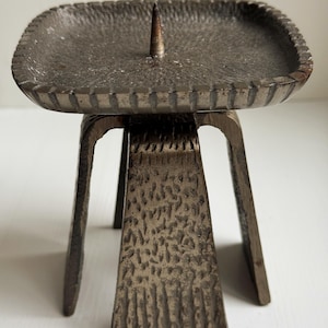 Puede incluir: Un candelero vintage de color bronce con una superficie texturizada. La parte superior cuadrada tiene un borde elevado y una punta central para una vela. El soporte está sostenido por una base de tres patas. Dimensiones: aprox. 10 cm x 10 cm.