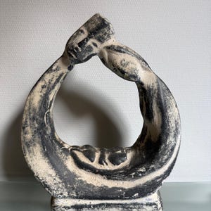 Op de afbeelding: Een crèmekleurig beeldhouwwerk met zwarte marmering. Het abstracte kunstwerk heeft een cirkelvormig ontwerp met twee figuren die elkaar omarmen. Het beeldhouwwerk is gemonteerd op een rechthoekige basis en is geschikt voor woondecoratie.