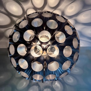 Op de afbeelding: Een bolvormige, metalen lamp met een complex ontwerp. De lamp is opgebouwd uit talrijke cilindrische buizen die in een bolvorm zijn gerangschikt, waardoor een patroon van cirkelvormige openingen ontstaat. De lichtbron binnenin werpt ovale schaduwen.