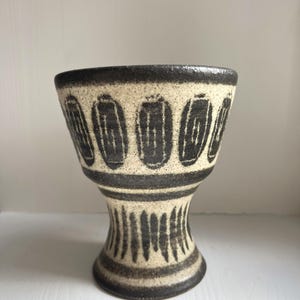Può includere: Un vaso in ceramica con base a piedistallo. Il vaso presenta un corpo color crema con motivi ovali marrone scuro attorno alla parte superiore e linee verticali sulla base. Il bordo e il bordo superiore sono marrone scuro.