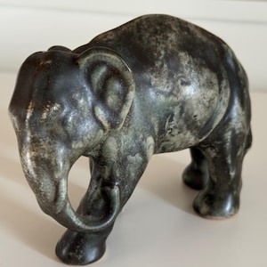 Peut inclure: Une figurine d'éléphant en céramique dans des tons de gris et de vert. L'éléphant est représenté en position de marche, sa trompe recourbée vers le bas. La figurine a une surface texturée et est un objet décoratif.
