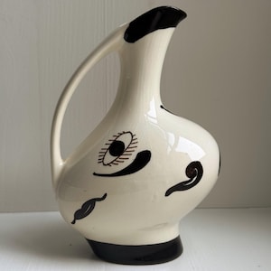 Op de afbeelding: Een vintage keramische kan met een crèmekleurige body en zwarte accenten. De kan heeft een gebogen handvat en een tuit met een zwarte rand. Decoratieve zwarte en bruine abstracte ontwerpen sieren de voorkant, waaronder een oogvorm en gebogen lijnen.