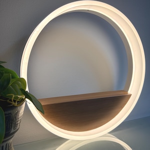 Peut inclure: Une étagère murale circulaire moderne avec une lumière LED intégrée. L'étagère présente un anneau blanc éclairé avec une étagère en bois. Le design est minimaliste et contemporain, adapté pour exposer des objets décoratifs ou des plantes. L'étagère mesure environ 30 cm de diamètre.