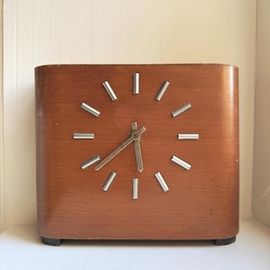 Könnte beinhalten: Eine rechteckige Holzuhr mit hellbraunem Finish. Das Zifferblatt hat silberfarbene Markierungen und Zeiger. Die Uhr ist im Vintage-Stil gehalten und steht auf einer weißen Oberfläche.