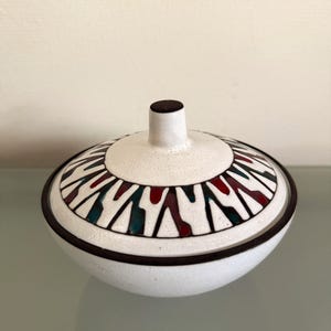 Op de afbeelding: Een ronde keramische kom met deksel in een matte witte afwerking. Het deksel heeft een centrale knop en een decoratieve band met herhalende geometrische vormen in donkerrood, blauwgroen en zwart. Een dunne zwarte lijn omlijnt de rand.