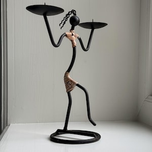 May include: Black metal candle holder in the shape of a stylized figure. The figure has a round head, two arms holding circular trays, and a wrapped waist. The base is a circular ring.
