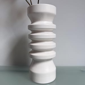 May include: A white ceramic vase with a unique, stacked design. The vase has a wide base and a narrower top, with several rounded sections in between. The vase holds a few stems with green leaves. The vase is a decorative home decor item.