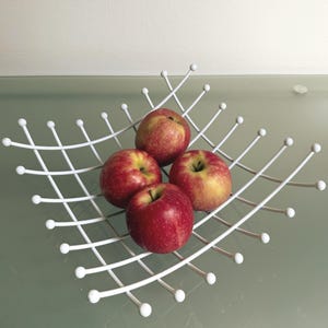 Op de afbeelding: Een witte draad fruitmand met vier rode appels met gele accenten. De mand heeft een geometrisch ontwerp met kleine witte bollen aan de uiteinden van de draden. De appels zijn rijp en vers.