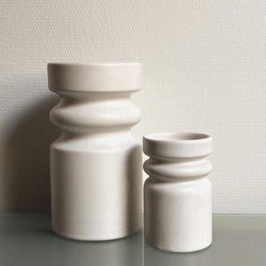 Op de afbeelding: Twee cilindrische, gebroken witte keramische vaten met een opvallend horizontaal ribbeldetail rond het bovenste gedeelte. Beide stukken hebben een subtiele craquelé glazuurafwerking, waarbij het ene vat merkbaar groter en breder is.