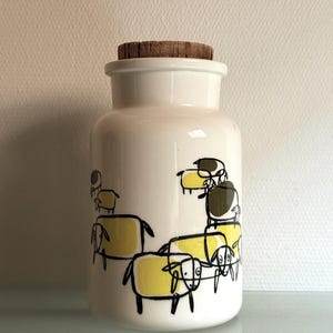 Op de afbeelding: Een cilindrische witte keramische voorraadpot met een kurken deksel. De buitenkant is voorzien van een herhalende zwarte lijntekening van schapen met gele en olijfgroene kleurvullingen. De pot heeft een gladde, glanzende afwerking en een brede basis.