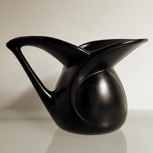 May include: A matte black ceramic pitcher featuring a smooth, rounded body and a wide, flared spout. The handle is integrated into the side, extending from the upper rim down to the base, creating a continuous, fluid silhouette with curved lines.